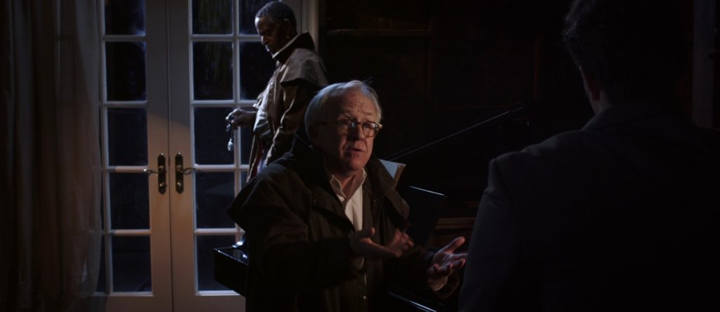 Scene from Until We Meet Again where Leslie Jordan is standing in front of Antonio Fargas annoyed that Jackson Rathbone's character isn't ready to move on. They are both wearing brown clothes and are in the piano room with a segmented window in the background.