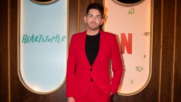 Scott McGlynn attends the Heartstopper event wearing a black jumper underneath a bright red suit.