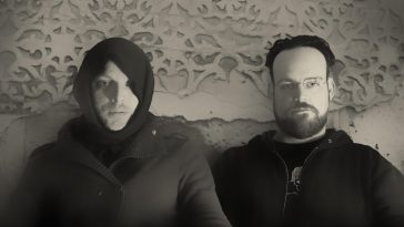 Black and white photo of The Drood with two guys centrallised on the picture, sitting down on a couch with the one on the left wearing a hoody with the hood up and the guy on the right wearing glasses and an open jacket.