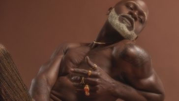 FEAT shows off his beautiful black skin in a confidence press picture for "This Time" which sees him posing topless with his head tilted to the right showing off his grey beard, and his left hand is in front of his body showcasing an assortment of rings and various gold jewellery pieces.
