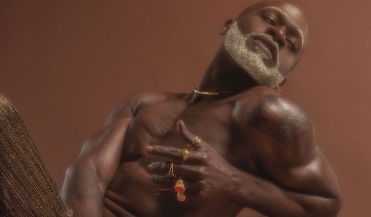 FEAT shows off his beautiful black skin in a confidence press picture for "This Time" which sees him posing topless with his head tilted to the right showing off his grey beard, and his left hand is in front of his body showcasing an assortment of rings and various gold jewellery pieces.