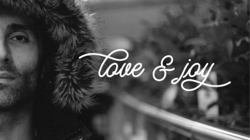 Black and white photo with The Guv on the left side of the photo showing only his left side of his face, and wearing a fur-lined coat with the hood up as he walks across a bridge. "Love and Joy" is written in white cursive font in the middle on the right side, and his name is written at the bottom. This is the single cover artwork.