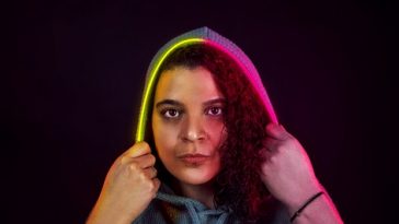 Studio promo photo of DJ G-String for "In The Mirror" which sees her standing in front of a black background with her putting an yellow-purple LED hood over her head.