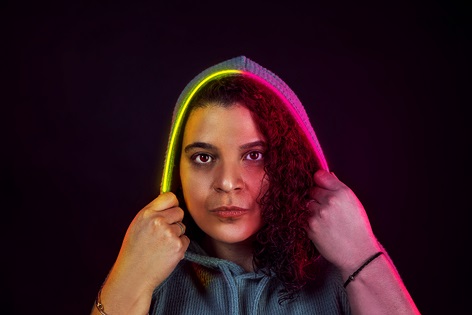 Studio promo photo of DJ G-String for "In The Mirror" which sees her standing in front of a black background with her putting an yellow-purple LED hood over her head.