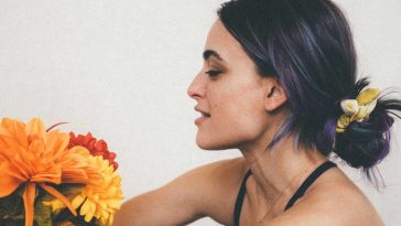 Grace Coletta in a promotional picture for "Daisies" sitting down facing to the left, with her purple-black hair bunched up in a bobble, and she's holding some sunflowers out towards her.