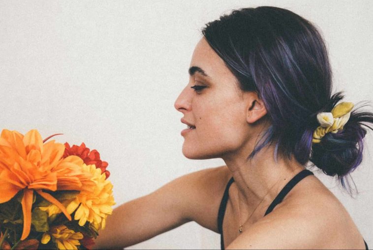 Grace Coletta in a promotional picture for "Daisies" sitting down facing to the left, with her purple-black hair bunched up in a bobble, and she's holding some sunflowers out towards her.