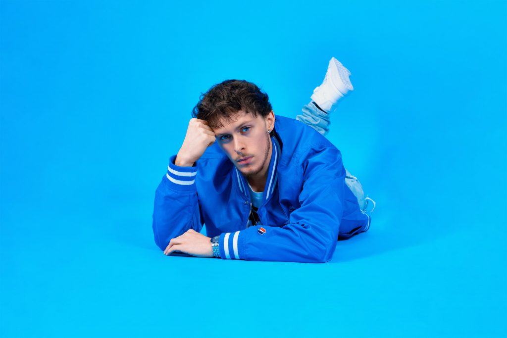 Scott the Pisces promoting "Summer '22" wearing a bright blue Superdry jacket, lying down on his front resting his head on his hand looking into the camera with his sultry blue eyes and curly brown hair, with one leg turned upwards at the back with a white trainer visible, posing in front of a bright blue background.