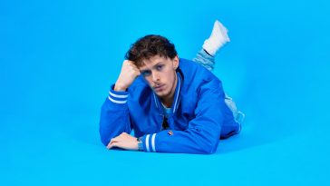 Scott the Pisces promoting "Summer '22" wearing a bright blue Superdry jacket, lying down on his front resting his head on his hand looking into the camera with his sultry blue eyes and curly brown hair, with one leg turned upwards at the back with a white trainer visible, posing in front of a bright blue background.