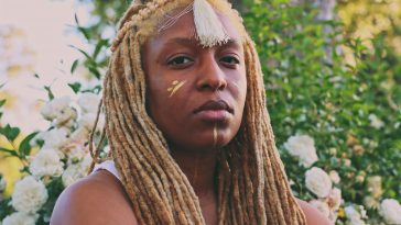 Promotional photo for ISSAMWERA with Yara Polana posing with blonde braids contrasting her black skin with tribal art on her face.
