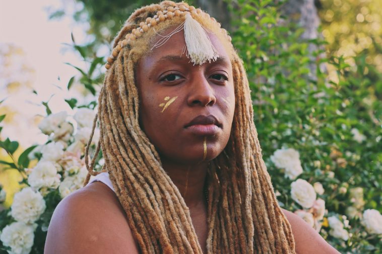 Promotional photo for ISSAMWERA with Yara Polana posing with blonde braids contrasting her black skin with tribal art on her face.