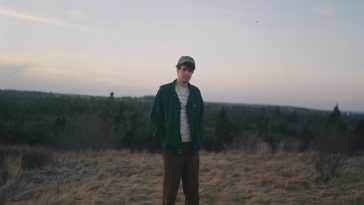 Promotional picture for "Oxford" album by Alan Hill which sees him standing in a field with his hands behind his back wearing a green jacket and brown chinos.