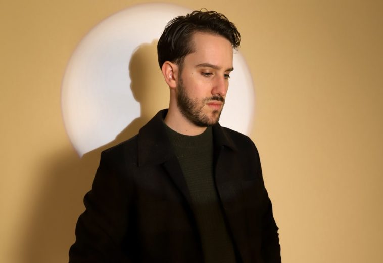 Promotional photo for "The Concept of You" by Ethan Mark which sees him in a spotlight against a beige background. He is wearing a black suit-jacket and a dark green jumper underneath. He is looking down towards his left.