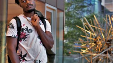 Gemïny posing in a press photo for "Too Close" which sees him outside with a glass building behind him. He has a white t-shirt with flowers on that contrasts his black skin, paired with jeans and a black rucksack. He's holding a jacket over one shoulder.