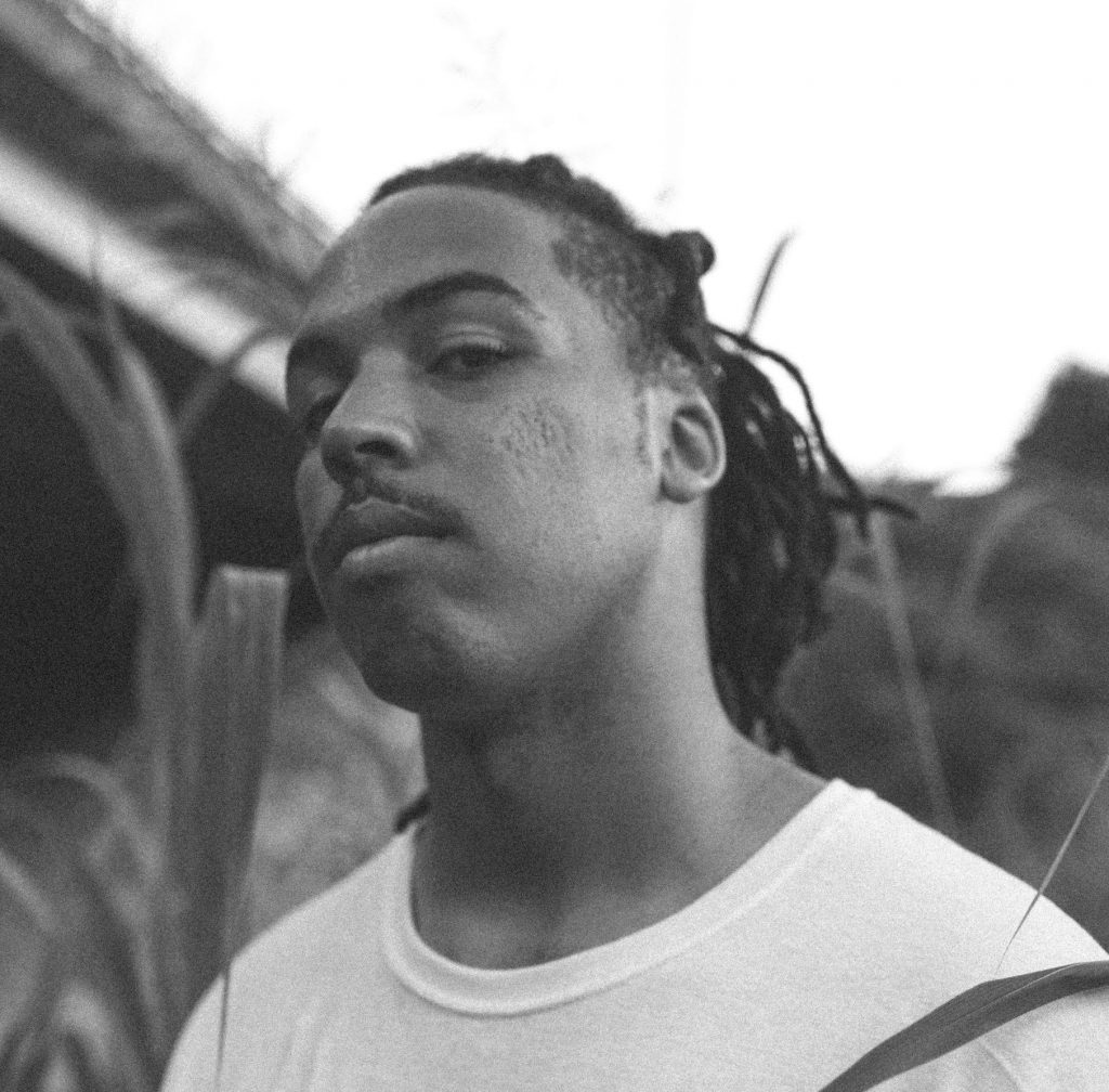Black and white promotional photo for "You're Not Gonna Like This" EP which is a close-up of Jeremiah Kingston who is wearing a white t-shirt and is looking down at the camera with a brigge in the background.