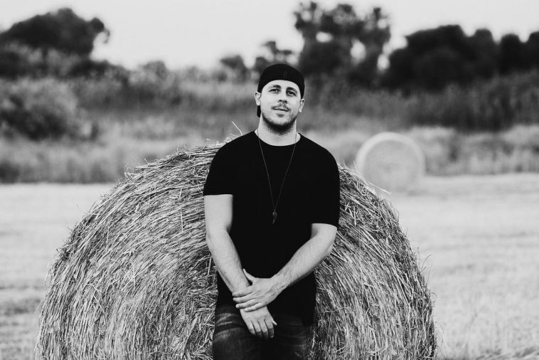 Singer Kais wearing a black t-shirt and a black snapback cap that he wears backwards, leaning against a haybale in the middle of a field in promotion for "Hot Air Balloon" EP.