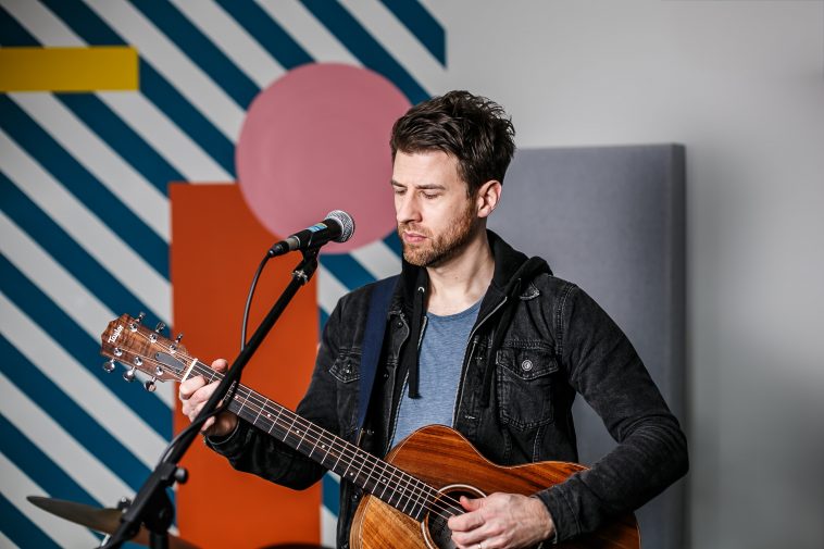 Promotional photo for "Rise" which sees Andy Hobson in a blue t-shirt under a black denim jacket, standing behind a microphone whilst holding a guitar, looking like he's about to perform. There's a diagonal blue and white striped wall behind him that has a pink circle and orange rectangle attached to it.