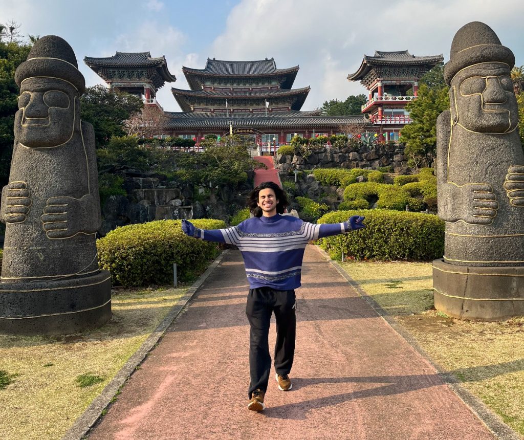 Promotional image for "Koryo Capsule" EP which sees Koosha Azim standing on a brick pave way with a temple behind him and two statue cylindrical monuments that have happy faces on and hats. Koosha Azim stands with his arms out wide, showing off his purple jumper and his black jeans, with a big smile on his face as his black hair hang around his shoulders.
