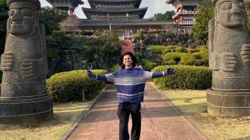 Promotional image for "Koryo Capsule" EP which sees Koosha Azim standing on a brick pave way with a temple behind him and two statue cylindrical monuments that have happy faces on and hats. Koosha Azim stands with his arms out wide, showing off his purple jumper and his black jeans, with a big smile on his face as his black hair hang around his shoulders.