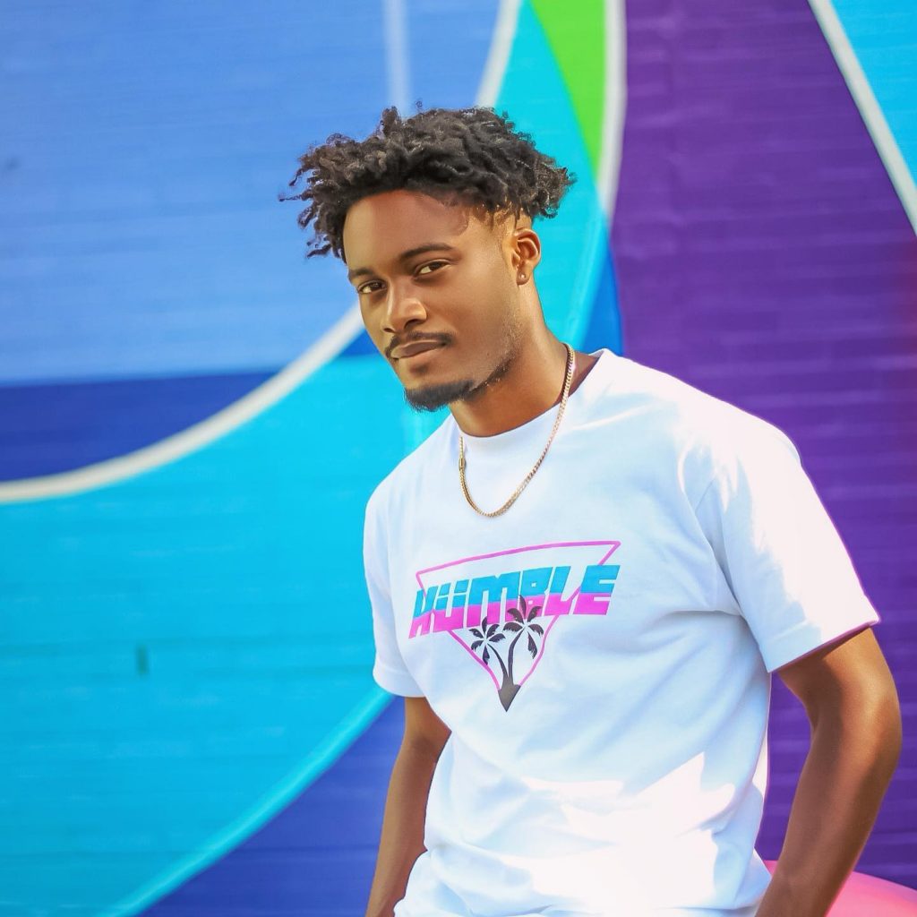 Promotional picture for "Hesitation" by Gemïny which sees him standing in front of a colourful swirl wall featuring blues, greens and purples, whilst wearing a white t-shirt that says "Humble" on the front in blue and pink lettering, paired with a gold chain necklace.
