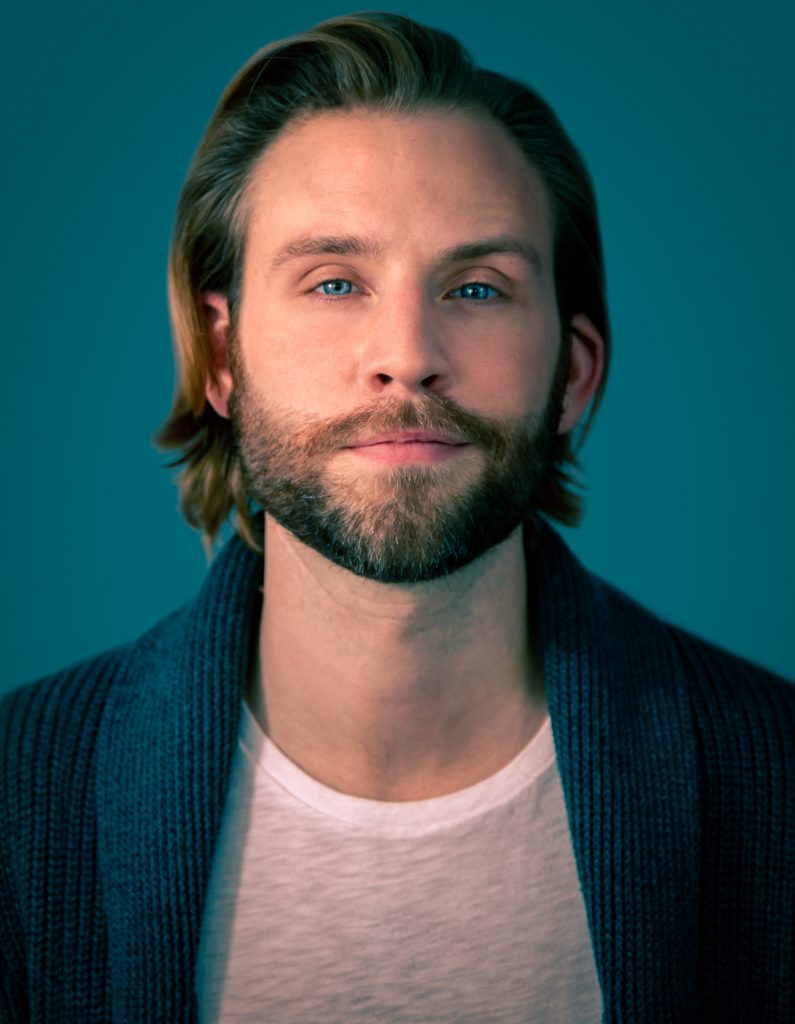 Headshot of Jeff Ayers for the NYC Web Fest 2022 which sees him with slicked back hair, wearing a white t-shirt under a blue blazer standing in front of a teal blue background.