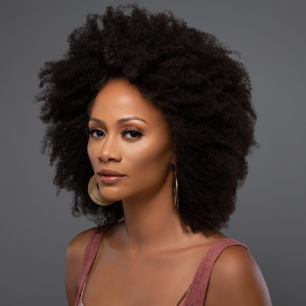Headshot of Nika King which sees her standing slightly to her right with her afro black hair perfect and wearing a pale pink strap top standing in front of a grey background.