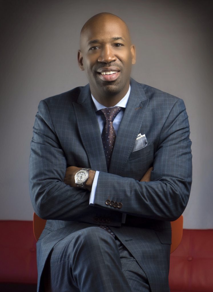 Heashot of J. Alexander Martin which sees him smiling whilst wearing a light navy blue suit with his arms crossed while leaning against a red backing in front of a grey backdrop.