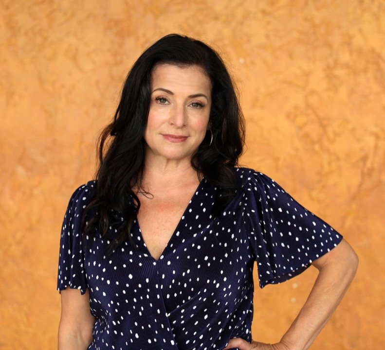 Headshot of Ria Pavia for the NYC Web Fest 2022 which sees her long black hair over both her shoulders and she's wearing a blue v neck blouse with white polka dots with her left hand on her hip. She's standing in front of an orange backdrop.