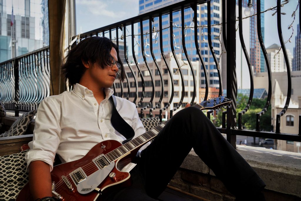 Promotional photo for "Relapse" which sees Vince Spano, wearing a white shirt and black trousers, sitting against a window, leaning his left side of his body against a black railing overlooking a cityscape. His left knee is raised slightly, his black hair is hiding the right side of his face as he looks out at the view with his glasses. He has a guitar strapped around him.
