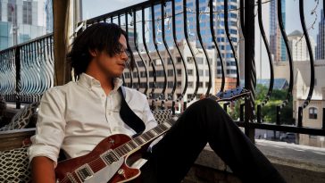 Promotional photo for "Relapse" which sees Vince Spano, wearing a white shirt and black trousers, sitting against a window, leaning his left side of his body against a black railing overlooking a cityscape. His left knee is raised slightly, his black hair is hiding the right side of his face as he looks out at the view with his glasses. He has a guitar strapped around him.