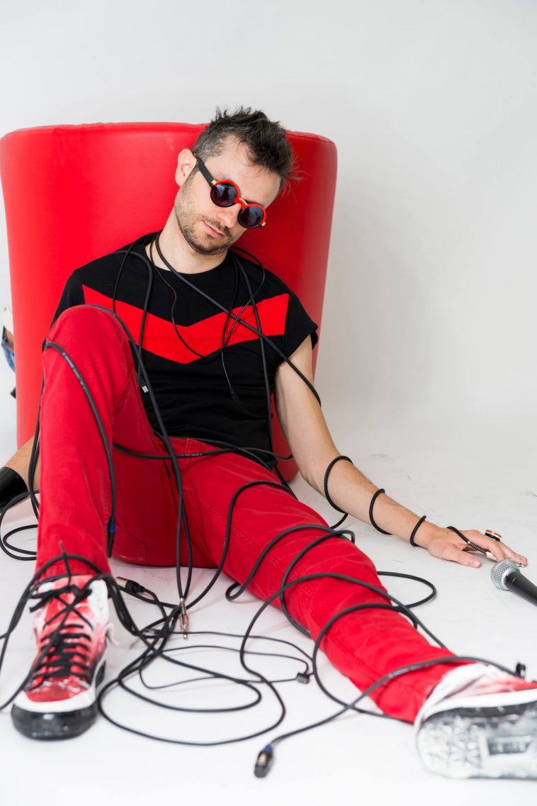 Promotional photo for "Run Coward" by Yarsha which sees him wearing red trousers and a black t-shirt with a red line across his chest, matched with black and red sunglasses, sitting on the floor with one knee up and the other leg straight, resting his back against a red cylindrical seat, wrapped up in a mess of wires.
