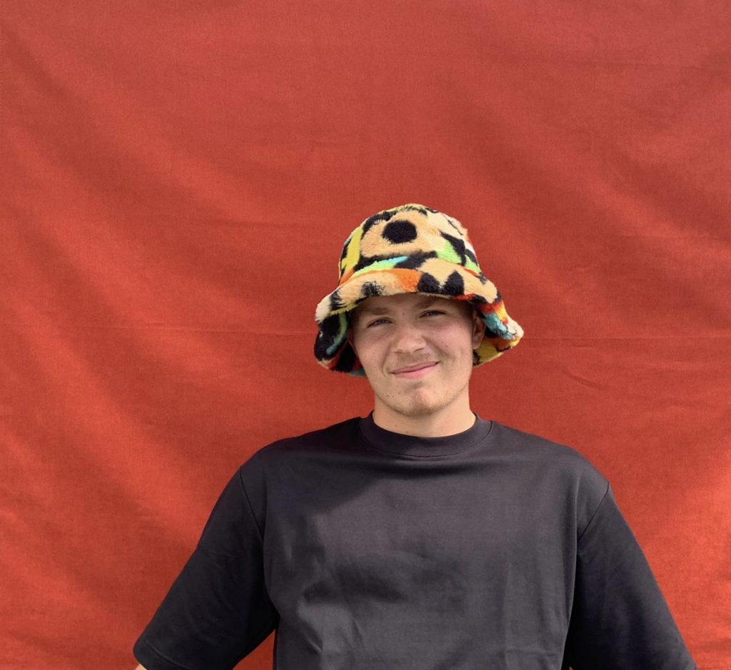 Promotional photo for "Someday" which sees Bradley Foster standing in front of a red background, wearing a long-sleeved t-shirt and a bright yellow with blue polka dots hat.