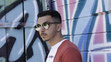 Promotional image for "Differences" which sees EM$o standing in front of a graffiti wall, facing to the left with sunglasses on and a red jacket over a white t-shirt.