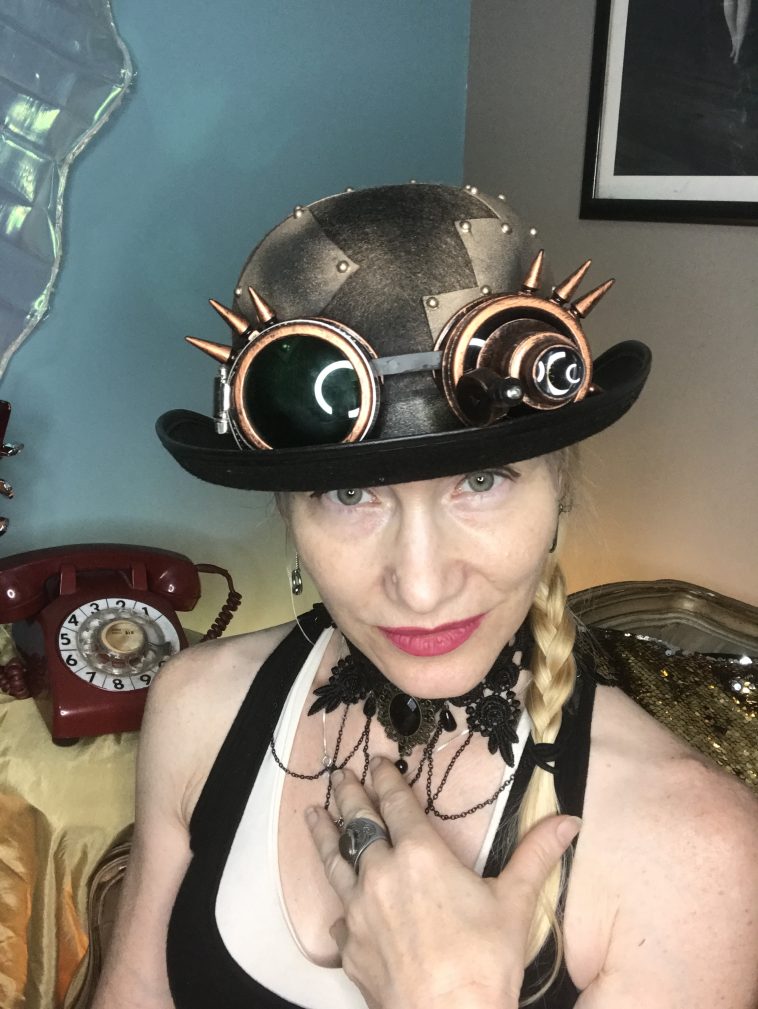 Promotional photo for "Reclaim Your Power" which sees Madam Who? taking a selfie wearing a steampunk hat with her blonde braid coming down her left shoulder, standing in front of an old red dial telephone and a blue background.