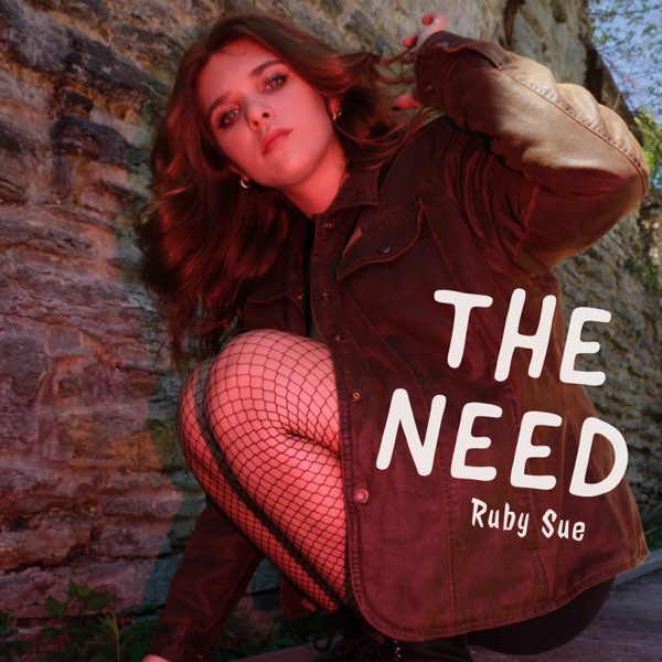Album cover artwork for "The Need" by Ruby Sue which sees her crouching with her right body against a brick wall, ,with her red coat on but still looking very xovers