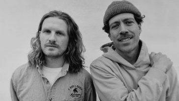 Black and white promotional photo for "Songs For The Weekend" which sees FRENSHIP standing next to one another. The guy on the left has shoulder-length hair and wearing a buttoned-up shirt over a tee, and the guy on the right is wearing a beanie hat and a hoodie - he also has his right arm draped across his chest with his hand on his left shoulder.