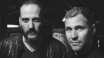 Promotional photo for "World Is Ours" in black and white which sees a headshot of the duo Ampersounds wearing leather jackets and looking into the camera, one of them has a beard and a short amount of hair on top, whereas the other one has a little bit of length to his hair and is clean-shaven.