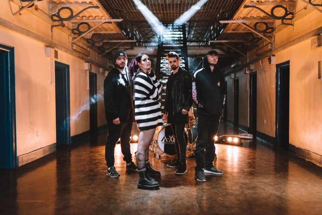 Promotional photo for "Mayday" which sees As December Falls posing on the ground floor of the prison where the music video is set. Bethany is in the front wearing a black and white striped oversized jumper, and her band mates are to the left and right of her wearing black.
