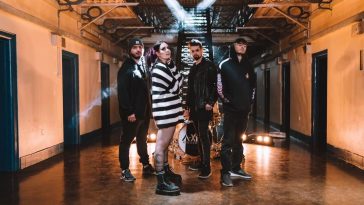 Promotional photo for "Mayday" which sees As December Falls posing on the ground floor of the prison where the music video is set. Bethany is in the front wearing a black and white striped oversized jumper, and her band mates are to the left and right of her wearing black.