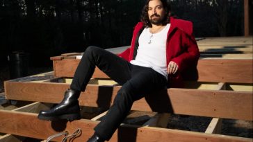 Promotional photo for "Ms Georgia" that sees Davey Harris relaxing on wooden bleachers in a forest. He is wearing a bright red jacket over a white v-neck t-shirt with black skinny jeans and black boots. His brown hair is shoulder-length and he has a prominent beard.