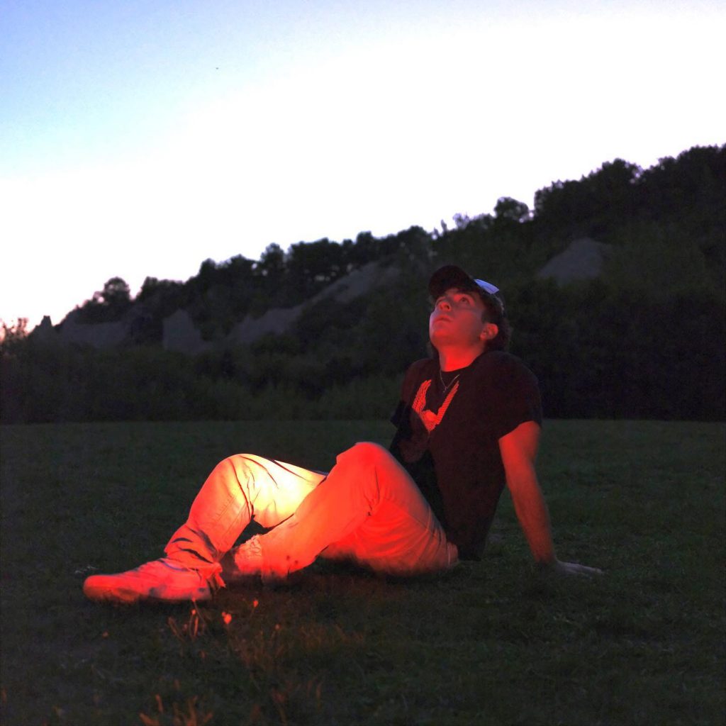 Promo picture for "September which sees Bye Alex sitting down in a field with a forest-like background and a blue and cloudy sky. He is wearing cream tracksuit bottoms, white trainers and a black t-shirt.