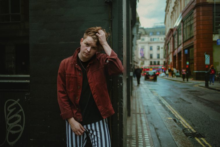 Promotional image for "All My Friends" which sees Drew Thomas wearing a red denim jacket over a black t-shirt with white and black striped trousers, posing with his left hand through his brown hair, on a side-street with the road going up his right. He is leaning against a black-painted building.