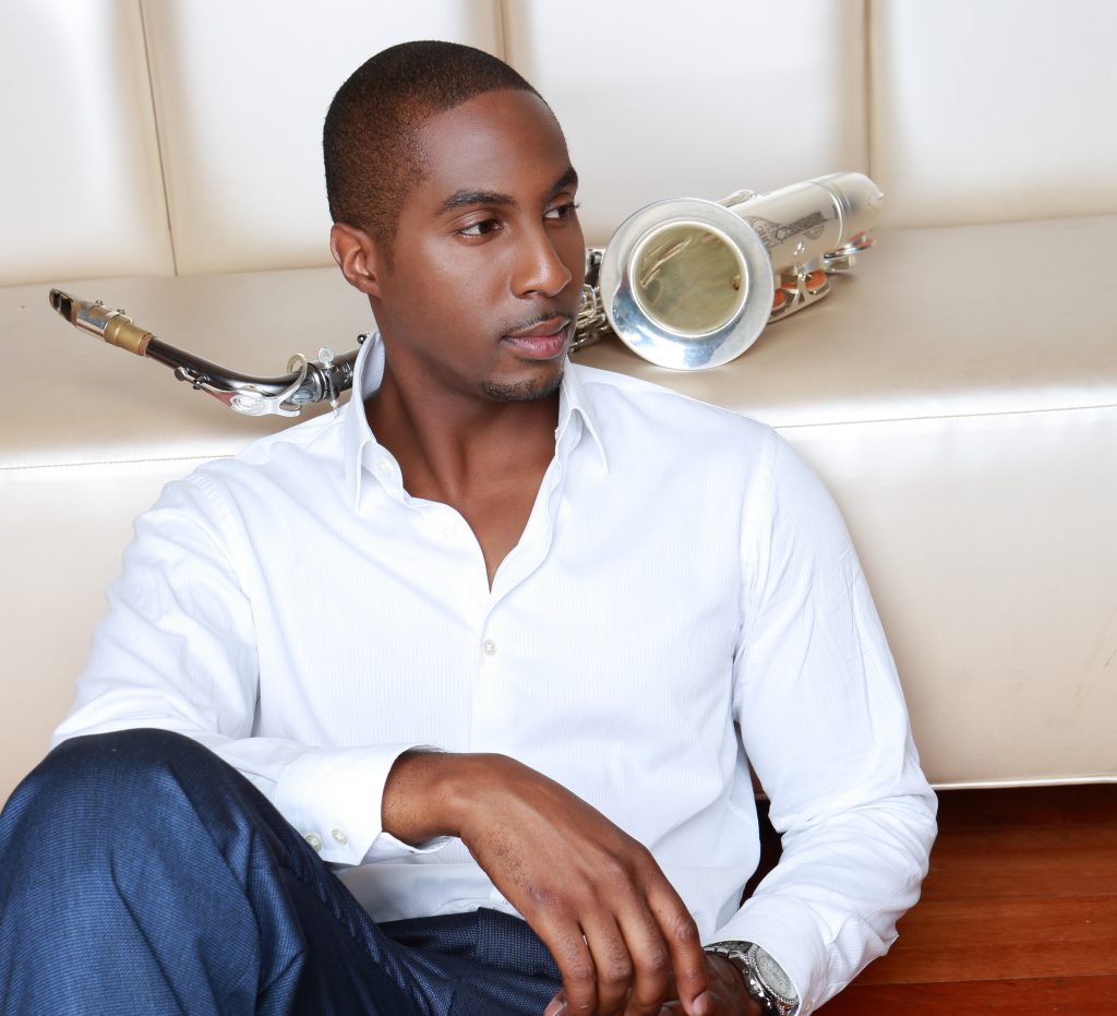 Eric Darius poses in a white shirt and blue jeans, sitting on the floor, resting his back against a white leather bench that has his saxophone lying on top, in promotion for "Unleashed".