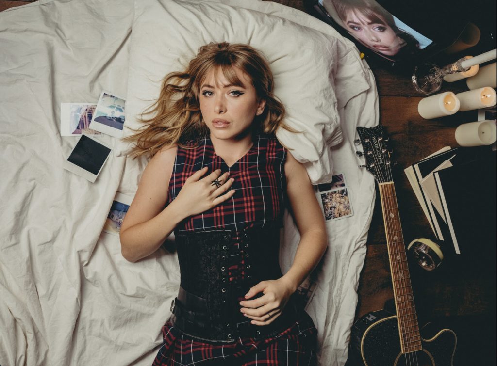 Promotional photo for "Before It Falls Apart" which sees Hunter Daily wearing a red and black checked dress with a black corset worn over the top, as she lies down on her back on her bed with some polaroid photos to her right and a guitar to her left. Her golden blonde hair is loosely spread in curls around her shoulders.