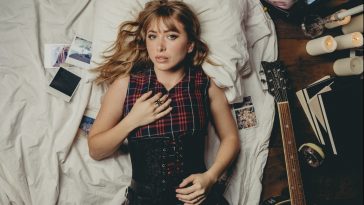 Promotional photo for "Before It Falls Apart" which sees Hunter Daily wearing a red and black checked dress with a black corset worn over the top, as she lies down on her back on her bed with some polaroid photos to her right and a guitar to her left. Her golden blonde hair is loosely spread in curls around her shoulders.