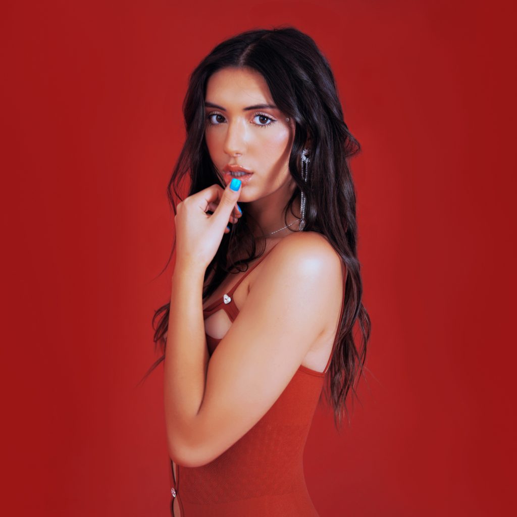 Promotional photo for "Heartbreaker" single which sees Juls posing with her thumb on her lips as she stares at the camera in a red halter dress, with a red background.