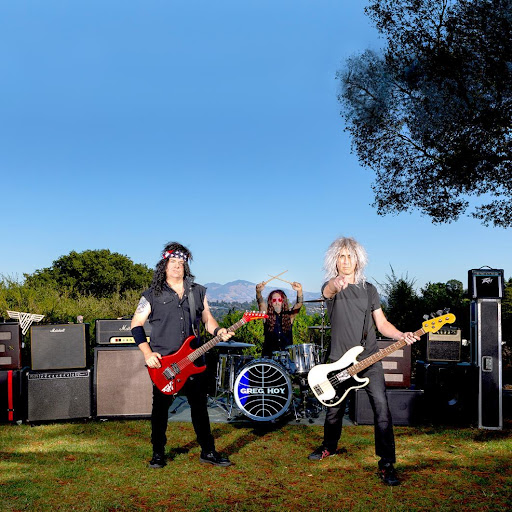 Promotional photo for "Demons At Night" which sees Greg Hoy & The Boys performing under a blue sky on a patch of grass with sound amps lining the back with the guys all wearing long-hair wigs reminiscent of the '80s with Greg Hoy and the guitarist up front holding their guitars and the drummer in the background.