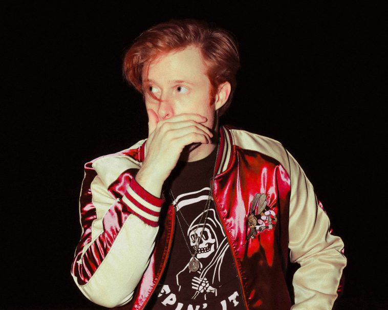 Promotional photo for "All I've Got To Say" which sees Jake Hays sitting down against a black background which illuminates his ginger hair which is short on the sides and a mid-length fringe. He is wearing a bright red and cream coloured bomber jacket over a black band t-shirt that has a white print on it. He has his right hand up to his mouth, almost with a shocked expression.