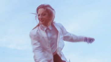 Promotional photo for "Your Heart Knows" which sees a low shot showing a bright blue sky with Jenny Kern running wearing a white jacket and matching top with short black shorts. The image has a blurred effect going on.