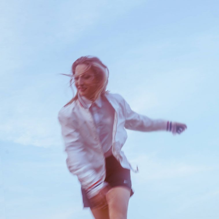 Promotional photo for "Your Heart Knows" which sees a low shot showing a bright blue sky with Jenny Kern running wearing a white jacket and matching top with short black shorts. The image has a blurred effect going on.