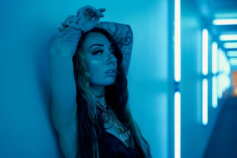 Promotional photo for "Drop The Top" which sees Kleøpatra leaning against a wall with her arms crossed on top of her head and she's looking sultry at the camera. She's got various necklaces around her neck and her black hair falls down her body and has two strips of blonde in front. The photo has been filtered with blue light.